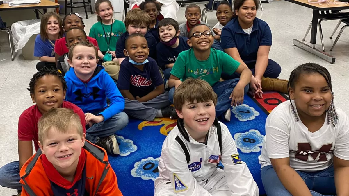 Students sit on a storytime carpet smiling and listening to their teacher (not pictured)