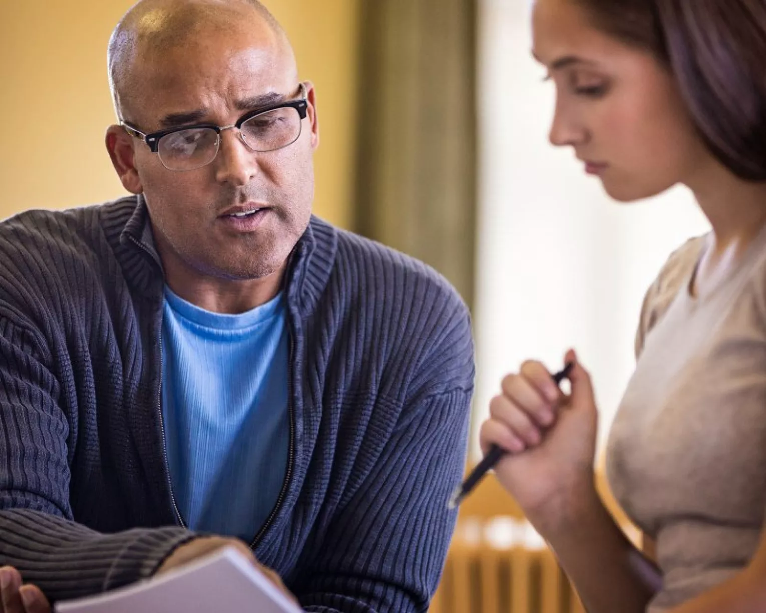 Two educators look over a checklist together. On the left, a bald man wearing glasses, a light blue t-shirt and a dark blue sweater holds a checklist in one hand and points to it with another. On the right, a woman in a tan colored shirt with long dark hair looks down at the list, holding a pen in her right hand.