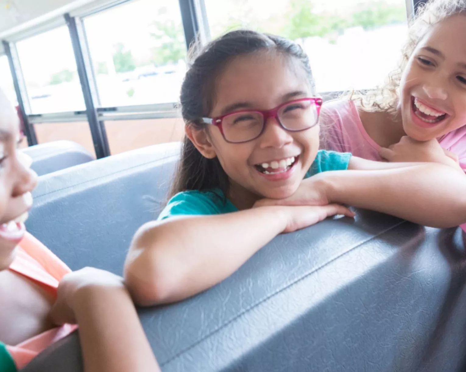 Three students sit on a bus leaning over a seat. All three are smiling.