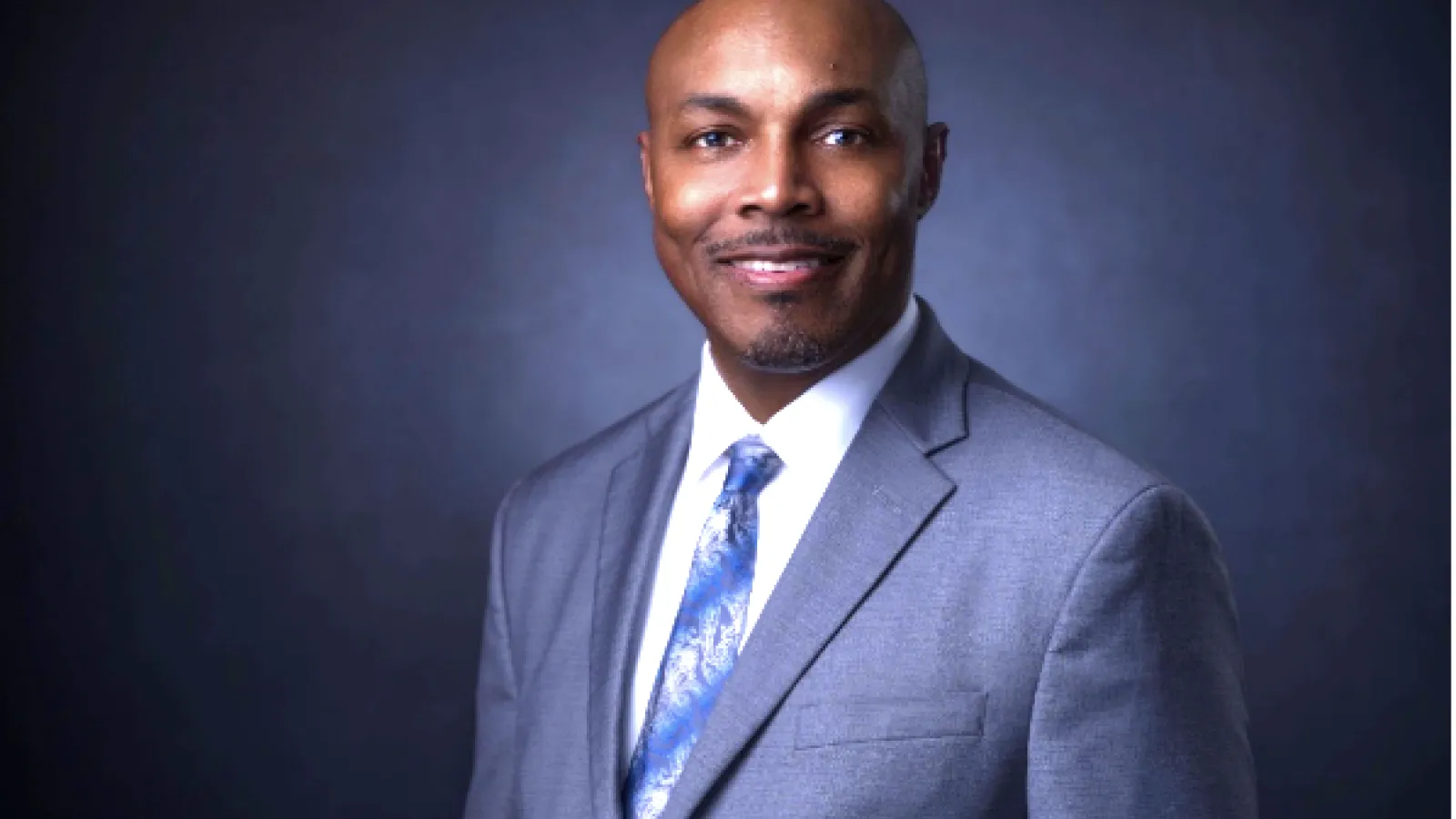 A professional headshot of a bald African American man wearing a gray suit, white dress shirt, and a patterned blue tie. He is smiling and looking directly at the camera against a dark gradient background.