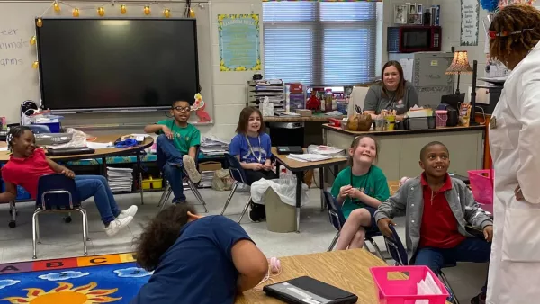 Several elementary school students sit in their desks looking at a classroom speaker in a white doctor's coat. The children are all smiling.