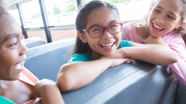 Three students sit on a bus leaning over a seat. All three are smiling.
