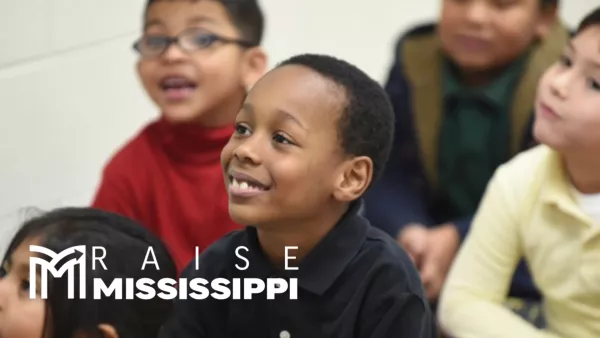 A group of students sit on a classroom rug for storytime. One student in the background in a red shirt with glasses is peeking at the camera with a mischevious smile. The camera is centered on a boy in a navy shirt with a slight smile as his eyes and attention are locked on the front of the classroom. In the corner, the Raise Mississippi logo appears in all white.