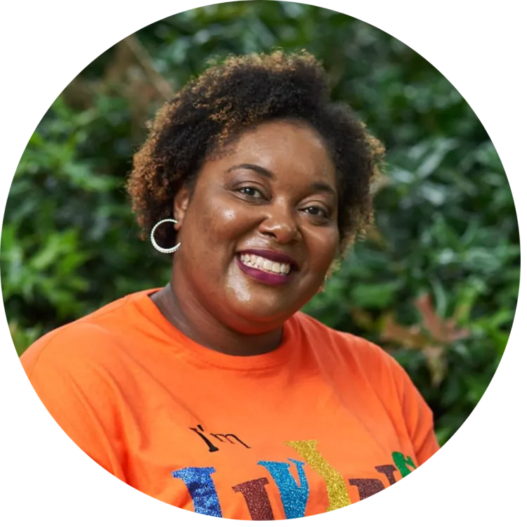 Erica Avent is in an orange shirt that says "I'm Living" in multi-color letters. She is wearing hoop earrings and smiling into the camera.